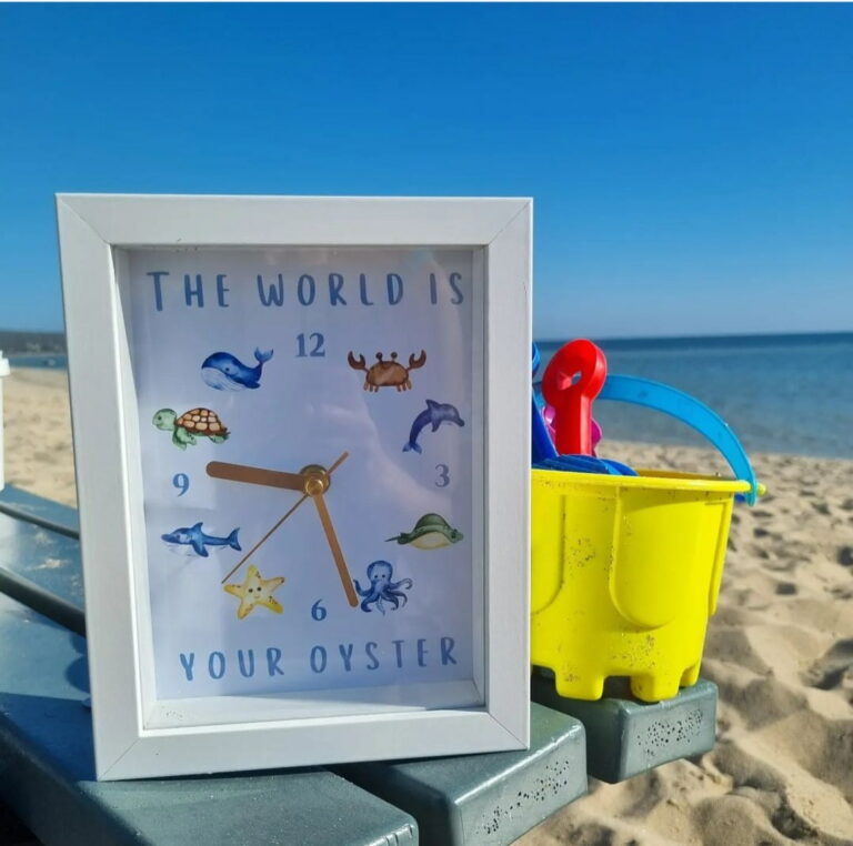 Custom made sea creatures picture clock sitting on bench in front of sandcastle bucket and shovel on the beach